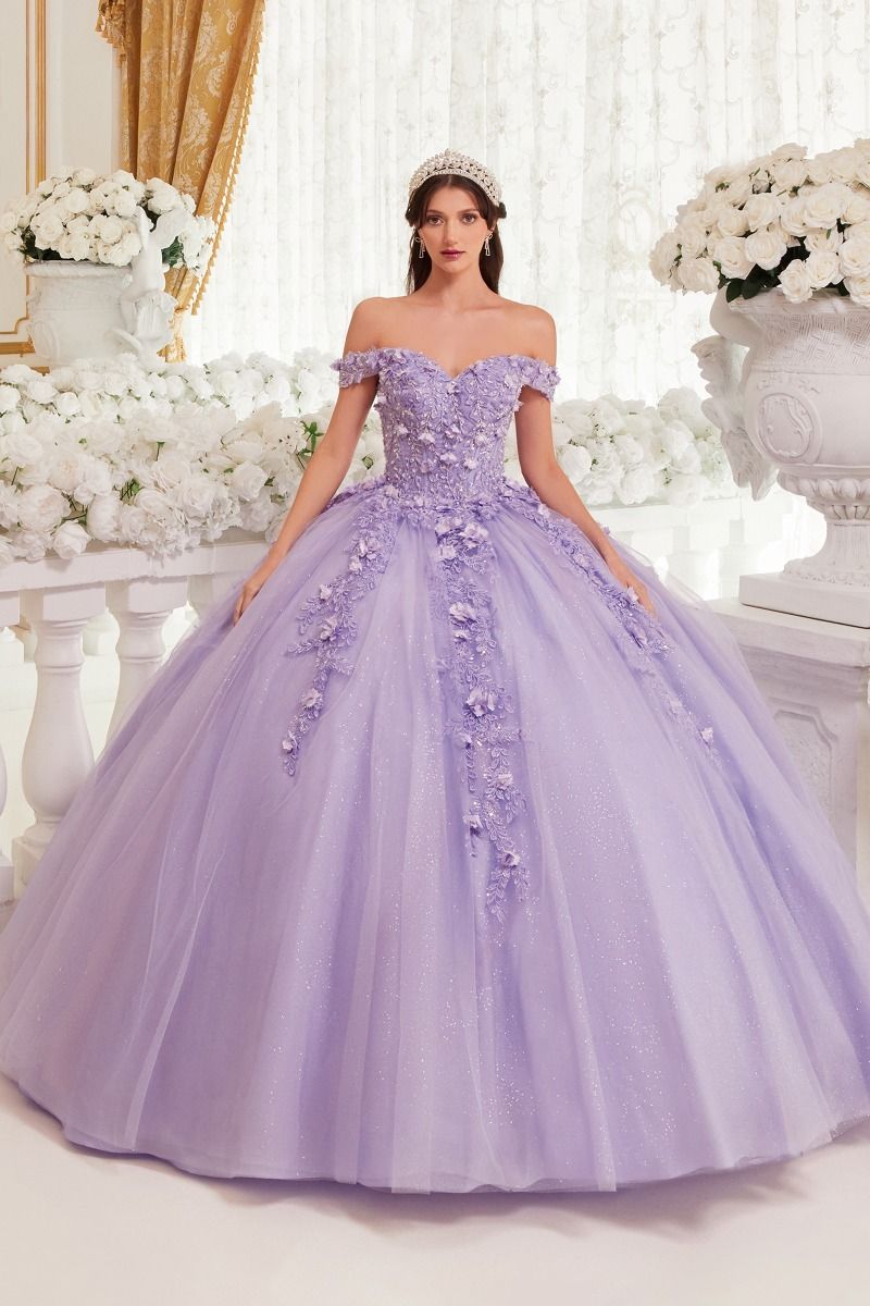 A woman stands indoors in the Off-Shoulder Purple Quinceanera Dress: Ladivine 15717, its floral details and glitter fabric sparkling among white flowers and elegant décor.